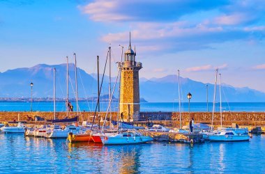The foggy evening on Lake Garda with yachts, Faro lighthouse and the silhouettes of Garda Prealps in background, Desenzano del Garda, Lombardy, Italy