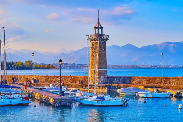 Historic stone lighthouse on Diga Foranea breakwater with yachts and boats in port and the Garda Prealps in the background at foggy sunset, Desenzano del Garda, Lake Garda, Italy