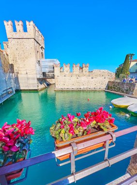The bridge of Sirmione marina with red cyclamens in pots with azure waters of Lake Garda and Scaligero Castle towers in background, Italy
