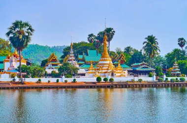 The hti umbrellas, chedi and spires of Wat Chong Klang Temple, surrounded with tropical garden in front of Nong Kham Lake, Mae Hong Son, Thailand
