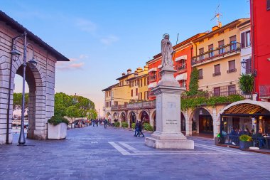 DESENZANO DEL GARDA - 10 Nisan 2022: Giuseppe Malvezzi Meydanı 'ndaki Sant' Angela anıtı 10 Nisan 'da Desenzano del Garda' da, renkli evler ve günbatımı gökyüzüne karşı