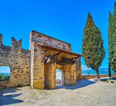 Yanı başında selvi ağaçları olan Brescia Kalesi 'nin taş kapısı ve arka planda Garda Prealps, İtalya
