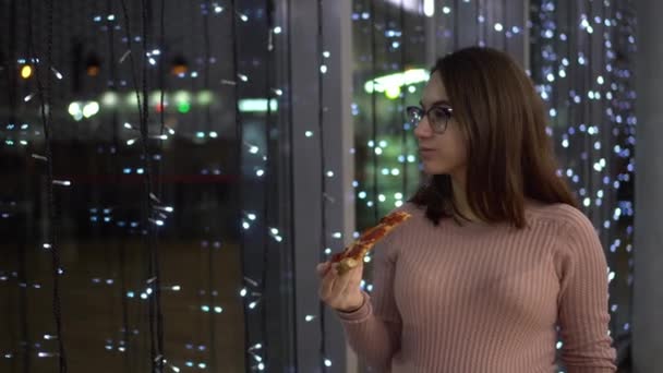Młoda kobieta w okularach je pizzę. Dziewczyna stoi przy panoramicznym oknie w centrum handlowym na tle girlandy. — Wideo stockowe