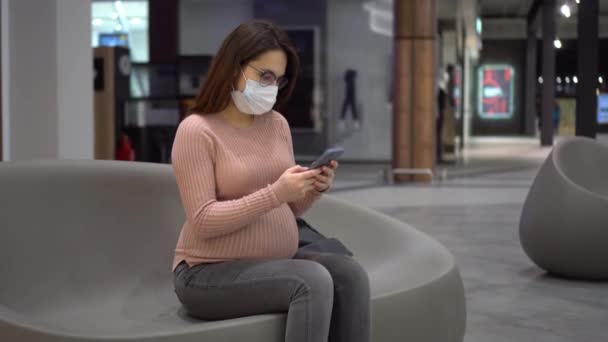 O tânără însărcinată cu ochelari și o mască medicală stă cu un telefon pe o bancă. Fata din mall și folosește un smartphone. — Videoclip de stoc