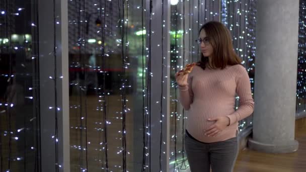 Een jonge zwangere vrouw met een bril die pizza eet. Een meisje staat bij het panoramische raam in een winkelcentrum tegen de achtergrond van slingers. — Stockvideo