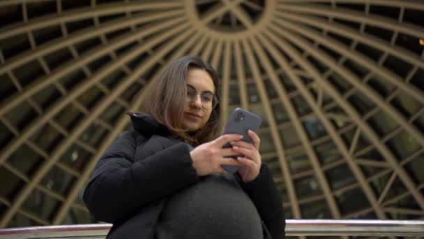 Una joven embarazada con gafas y una chaqueta de pie con un teléfono en las manos. Una chica en un centro comercial utiliza un teléfono inteligente contra el telón de fondo de una cúpula panorámica. — Vídeo de stock
