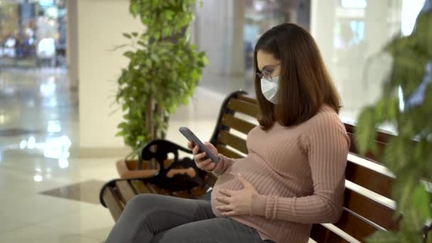 Uma jovem grávida de óculos e uma máscara médica senta-se com um telefone em um banco. Menina no shopping e usa um smartphone. — Vídeo de Stock