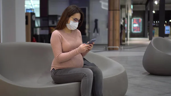 Μια νεαρή έγκυος γυναίκα με γυαλιά και μια ιατρική μάσκα κάθεται με ένα τηλέφωνο σε ένα παγκάκι. Κορίτσι στο εμπορικό κέντρο και χρησιμοποιεί ένα smartphone. — Φωτογραφία Αρχείου