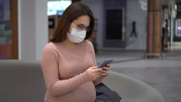 Μια νεαρή έγκυος γυναίκα με γυαλιά και μια ιατρική μάσκα κάθεται με ένα τηλέφωνο σε ένα παγκάκι. Κορίτσι στο εμπορικό κέντρο και χρησιμοποιεί ένα smartphone. — Φωτογραφία Αρχείου