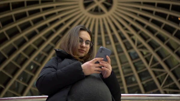 Egy fiatal terhes nő szemüvegben és egy kabát áll egy telefonnal a kezében. Egy lány a bevásárlóközpontban okostelefont használ egy panoráma kupola hátterében.. — Stock Fotó