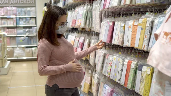 Mladá těhotná žena s brýlemi a lékařskou maskou v obchodě s dětmi. Dívka si vybírá oblečení pro nenarozené dítě. — Stock fotografie