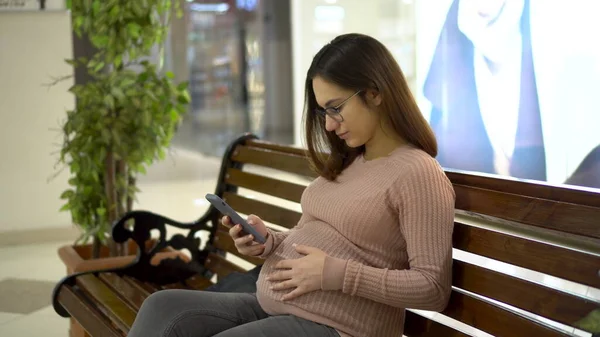 Uma jovem grávida de óculos senta-se com um telefone em um banco. Menina no shopping e usa um smartphone. — Fotografia de Stock