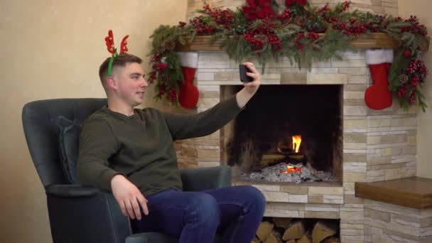 Młody mężczyzna siedzi w fotelu przy kominku z rogami na głowie i robi sobie selfie. Świąteczny nastrój. — Wideo stockowe