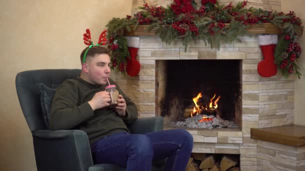 A young man sits in an armchair by the fireplace with horns on his head and drinks cocoa. Christmas mood. — Stock Video