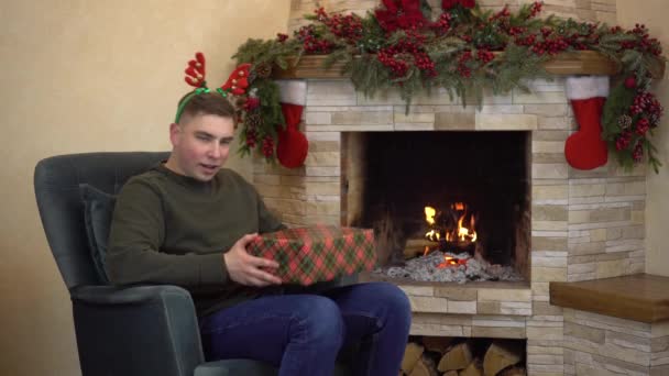 Een jongeman zit in een fauteuil bij de open haard met hoorns op zijn hoofd en houdt geschenken vast en schudt. Kerststemming. — Stockvideo