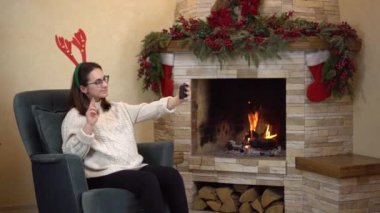 Gözlüklü hamile bir kadın şöminenin yanındaki koltukta oturur ve boynuzlarıyla selfie çeker. Noel havası.