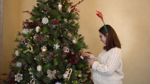 Eine junge schwangere Frau in Brille und Pullover mit Hörnern auf dem Kopf begutachtet das Spielzeug am Weihnachtsbaum. Weihnachtsstimmung. — Stockvideo