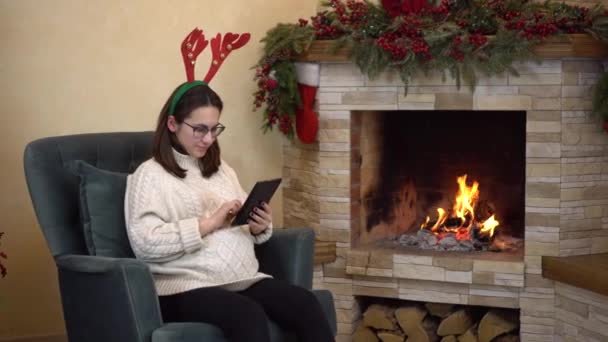 Een jonge zwangere vrouw met een bril zit in een stoel bij de open haard met horens op haar hoofd en schrijft op de tablet. — Stockvideo