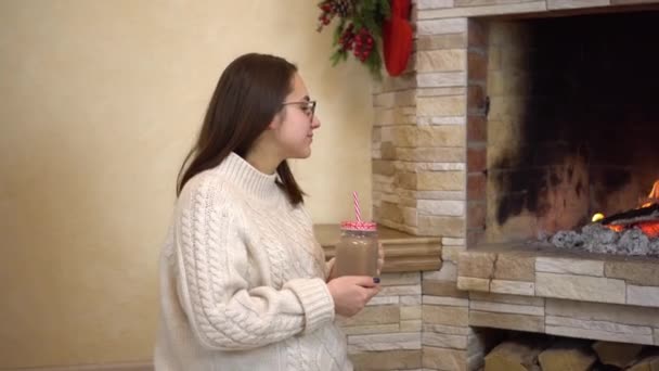 A young pregnant woman with glasses sits by the fireplace and drinks cocoa. Christmas mood. — Stock Video