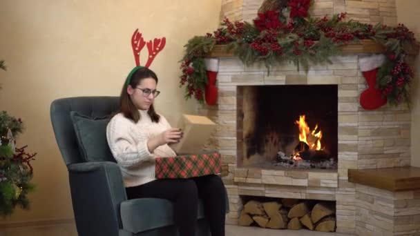 Een jonge zwangere vrouw met een bril zit in een stoel bij de open haard met horens op haar hoofd en houdt geschenken vast en schudt. Kerststemming. — Stockvideo