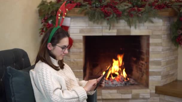 A young pregnant woman with glasses sits in a chair by the fireplace with horns on her head and writes on the tablet. — Stock Video