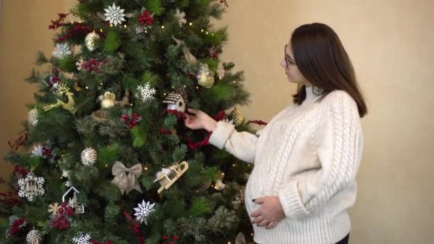 Een jonge zwangere vrouw met een bril en een trui onderzoekt speelgoed op de kerstboom. Kerststemming. — Stockvideo