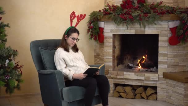Młoda ciężarna kobieta w okularach siedzi w fotelu przy kominku z rogami na głowie i czyta książkę. Świąteczny nastrój. — Wideo stockowe