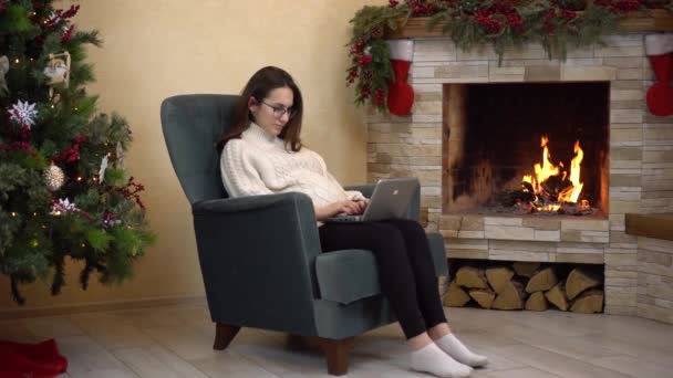 Una joven embarazada con gafas se sienta en un sillón junto a la chimenea y se imprime en un ordenador portátil. La chica escribe un correo electrónico a Santa Claus. — Vídeos de Stock