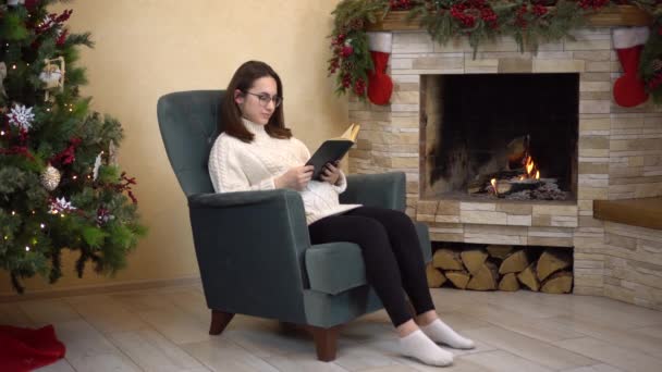 Een jonge zwangere vrouw met een bril zit in een fauteuil bij de open haard en leest een boek. Kerststemming. — Stockvideo