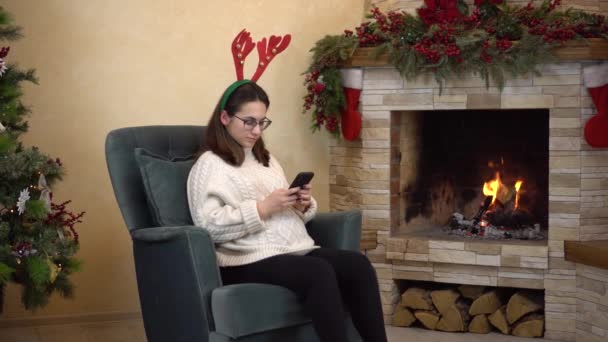 En ung gravid kvinna med glasögon sitter i en fåtölj vid öppna spisen med horn på huvudet och använder en smartphone. — Stockvideo
