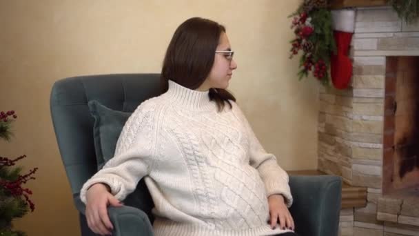 O tânără însărcinată cu ochelari într-un pulover stă într-un fotoliu lângă șemineu și se uită la foc. Moda Crăciunului. — Videoclip de stoc