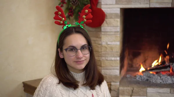 Una joven embarazada con gafas se sienta junto a la chimenea con cuernos en la cabeza y bebe cacao. Humor de Navidad. — Foto de Stock