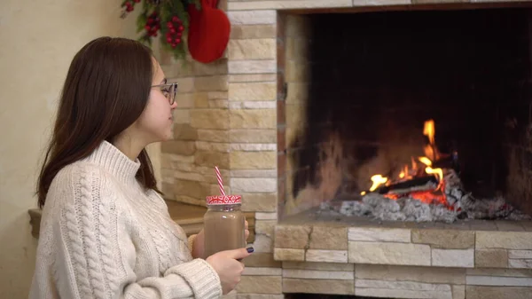 Μια νεαρή έγκυος γυναίκα με γυαλιά κάθεται δίπλα στο τζάκι και πίνει κακάο. Χριστουγεννιάτικη διάθεση. — Φωτογραφία Αρχείου