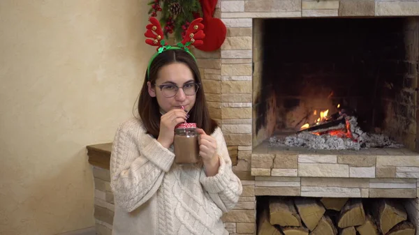 A young pregnant woman with glasses sits by the fireplace with horns on her head and drinks cocoa. Christmas mood. — Stock Photo, Image