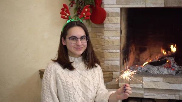 Una joven embarazada en gafas se sienta junto a la chimenea con un bengala en la mano. Humor de Navidad. —  Fotos de Stock