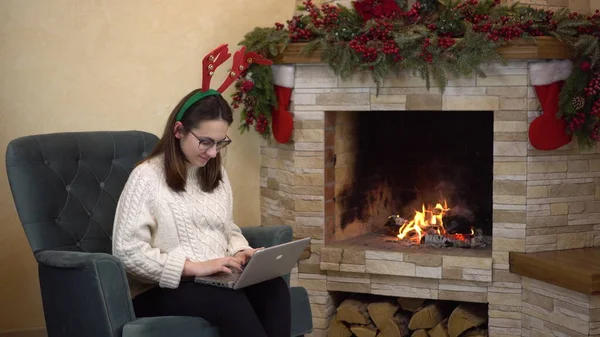 O tânără însărcinată cu ochelari stă într-un fotoliu lângă șemineu cu coarne pe cap și tastează pe un laptop. Fata îi scrie un e-mail lui Moş Crăciun. — Fotografie, imagine de stoc