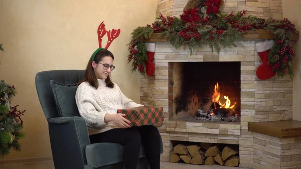 A young pregnant woman with glasses sits in a chair by the fireplace with horns on her head and holds and shakes gifts. Christmas mood. — Stock Photo, Image