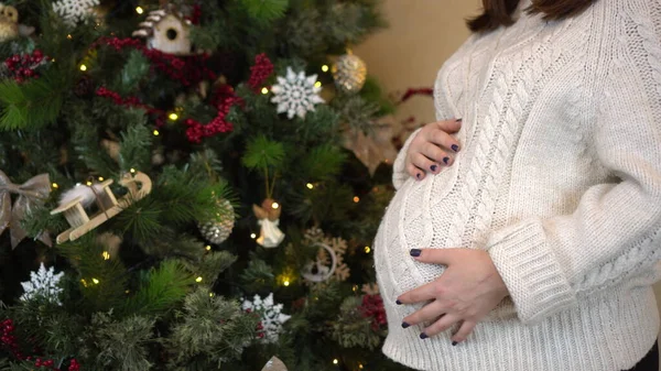 Mladá těhotná žena v bílém svetru drží břicho v blízkosti vánočního stromečku close-up. — Stock fotografie