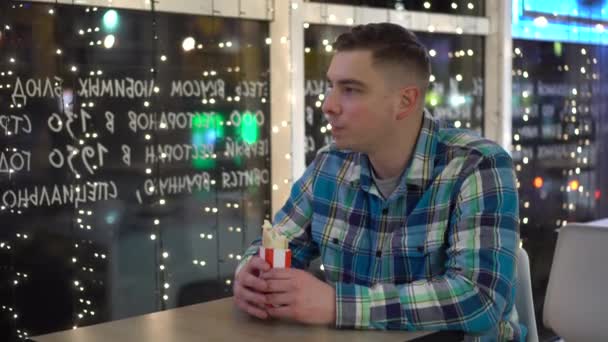 Un tânăr mănâncă burrito într-un restaurant. Tipul stă într-o cafenea seara lângă fereastra decorată cu ghirlande. — Videoclip de stoc