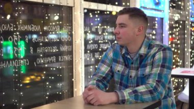 Genç bir adam bir kafede bir masada oturur ve pencereden dışarı bakar. Adam akşam üstü pencerenin yanındaki bir restoranda çelenklerle süslenmiş bir şekilde oturuyor..