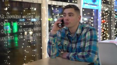 Genç bir adam akşam bir kafede bir masada oturur ve telefonda konuşur. Çelenklerin arkasında akıllı telefonu olan bir adam..
