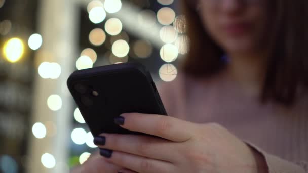 Uma jovem está sentada em uma mesa em um café à noite e mensagens de texto ao telefone. Menina com um telefone nas mãos no fundo de guirlandas. Close-up. — Vídeo de Stock