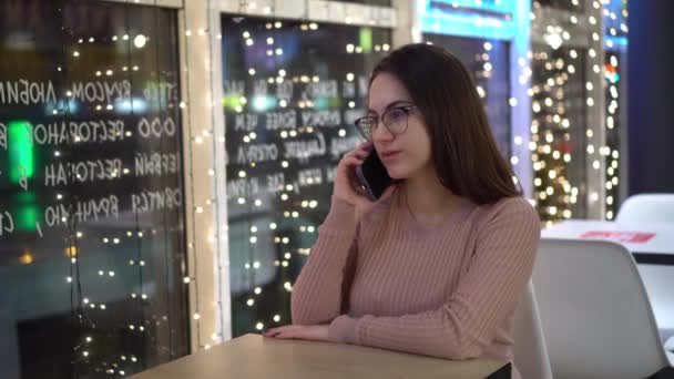 Uma jovem senta-se a uma mesa em um café à noite e fala ao telefone. Menina com um smartphone no fundo de guirlandas. — Vídeo de Stock