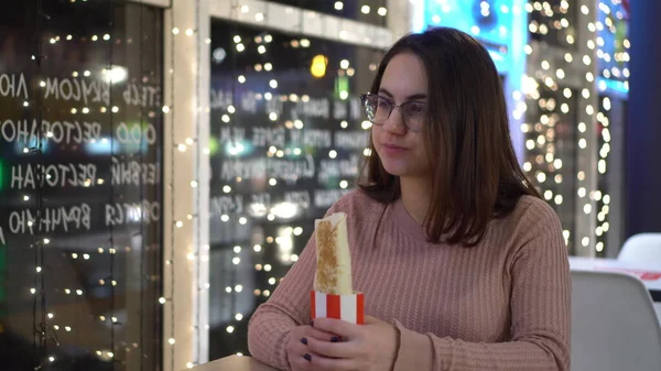 Jeune femme à lunettes mangeant du burrito dans un restaurant. Une fille s'assoit dans un café le soir près de la fenêtre décorée de guirlandes. — Photo