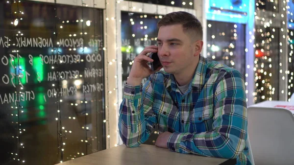 Вечером молодой человек сидит за столом в кафе и разговаривает по телефону. Человек со смартфоном на фоне гирлянд. — стоковое фото