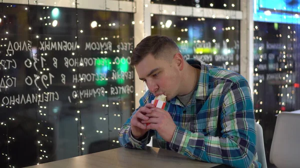 Jovem a comer burrito num restaurante. O cara se senta em um café à noite pela janela decorada com guirlandas. — Fotografia de Stock