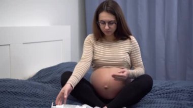 Young pregnant woman with glasses smears cream on her belly. Girl in the bedroom on the bed.
