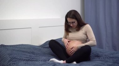 Young pregnant woman with glasses smears cream on her belly. Girl in the bedroom on the bed.