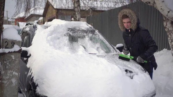一个穿着夹克的年轻人用刷子擦雪地.汽车被雪覆盖着.天气灾难. 图库照片