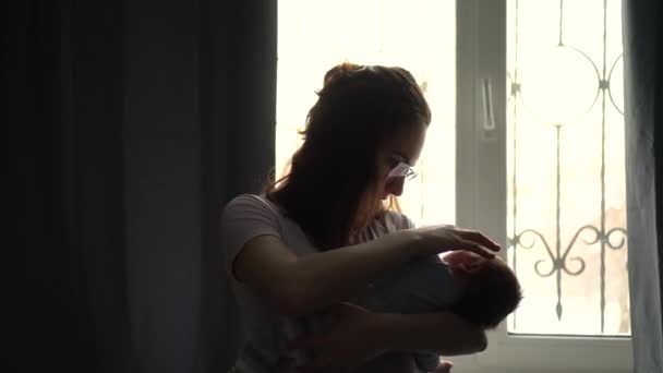 O tânără mamă ține un copil în brațe pe fundalul unei ferestre. Lumina de la fereastră luminează imaginea unei femei cu un nou-născut. — Videoclip de stoc